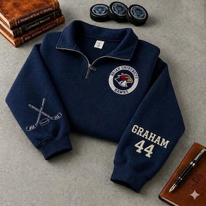 Peut inclure: Un sweat-shirt bleu marine à fermeture éclair quart, arborant le logo Briar University Hawks sur la poitrine, des crosses de hockey croisées sur la manche droite et le nom GRAHAM avec le numéro 44 brodés en blanc sur la manche gauche.