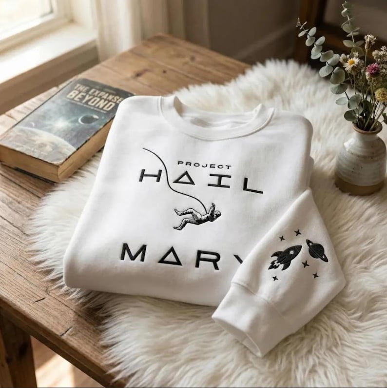 May include: White sweatshirt with the text "PROJECT HAIL MARY" in black, featuring an astronaut graphic. The sleeve displays a rocket and planet design. A book titled "THE EXPANSE BEYOND" rests on a wooden surface, alongside a vase of flowers.
