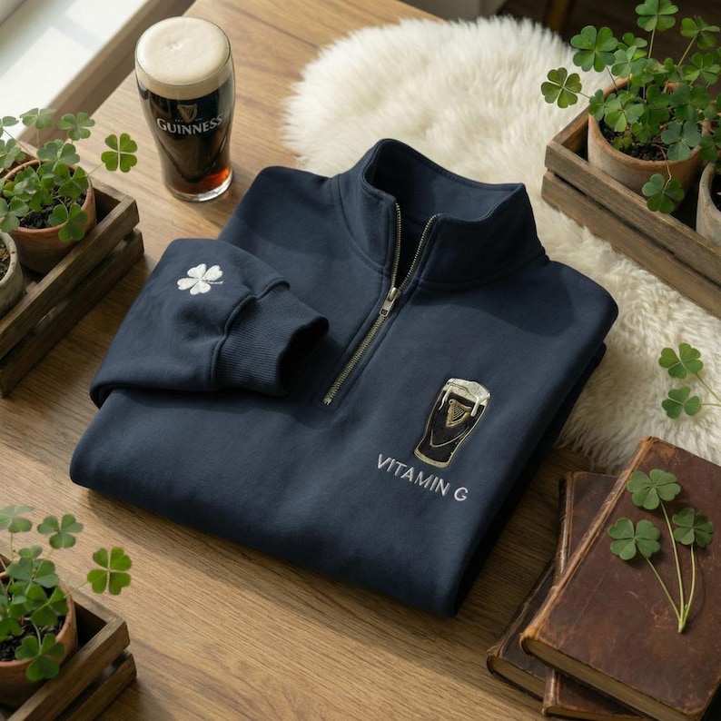 Pode incluir: Camisola azul marinho com fecho de correr de um quarto, fecho de correr dourado e desenhos bordados de trevos e copos de Guinness. O texto "Vitamin G" est&aacute; abaixo. Um copo de Guinness &eacute; vis&iacute;vel.