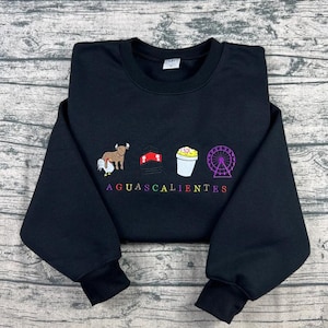 May include: Black sweatshirt featuring embroidered icons of a bull, a hat, a cup of ice cream, and a Ferris wheel. The word "AGUASCALIENTES" is embroidered in rainbow colours. The sweatshirt has a crew neck and long sleeves.