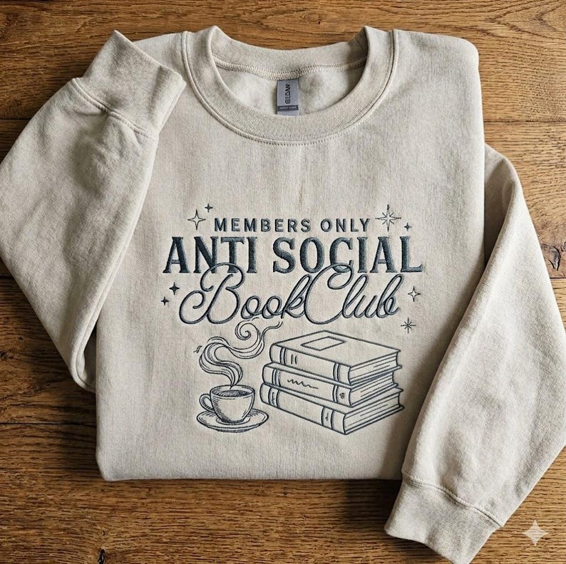 May include: A beige crewneck sweatshirt featuring dark gray embroidery. The text reads "MEMBERS ONLY ANTI SOCIAL Book Club" above a stack of three books and a steaming cup of coffee. Small star icons are scattered around the central design.