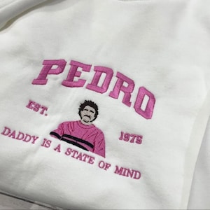 Peut inclure: Sweat-shirt blanc avec broderie rose. Le mot "PEDRO" est brodé en arc de cercle. En dessous, le texte "EST. DADDY IS A STATE OF MIND 1975" et un dessin d'un homme en chemise rose.