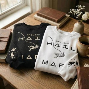 May include: Two sweatshirts, one black and one white, with the text "PROJECT HAIL MAF" and an astronaut graphic. The black sweatshirt has a rocket and planet design on the sleeve. Both are on a wooden table.