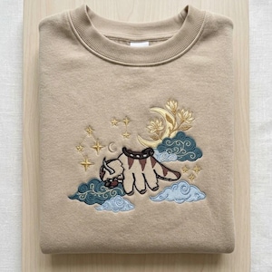 May include: A beige crewneck sweatshirt featuring an embroidered design of a flying bison creature with brown markings. The embroidery includes a yellow crescent moon, stylized floral elements, dark teal and light blue clouds, and gold stars.