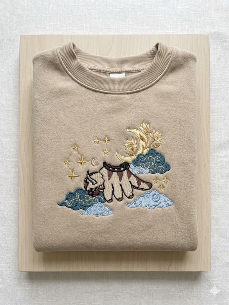 May include: A beige crewneck sweatshirt featuring an embroidered design of a flying bison creature with brown markings. The embroidery includes a yellow crescent moon, stylized floral elements, dark teal and light blue clouds, and gold stars.