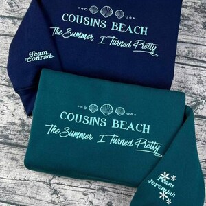 Peut inclure: Deux sweat-shirts, un bleu marine et un vert d'eau, avec du texte blanc. Le texte sur le devant indique "COUSINS BEACH The Summer I Turned Pretty". Le sweat-shirt bleu marine porte "Team Conrad" sur la manche, et le vert d'eau "Team Jeremiah".