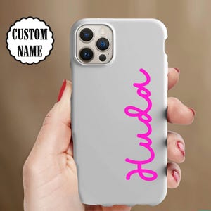 May include: A light gray phone case with the name "Huda" in bright pink cursive script. The phone case is held in a hand with red nail polish. The top left corner of the image shows a white and black circular graphic that says "CUSTOM NAME".