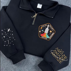Könnte beinhalten: Schwarzes Sweatshirt mit goldenem Reißverschluss und gestickten Aufnähern. Ein Aufnäher zeigt eine Rakete mit dem Text "HAIL MARY ИАИСКИHA GRACE". Der andere Ärmel hat das Wort "AMAZE" wiederholt. Eine Konstellation befindet sich auf dem anderen Ärmel.