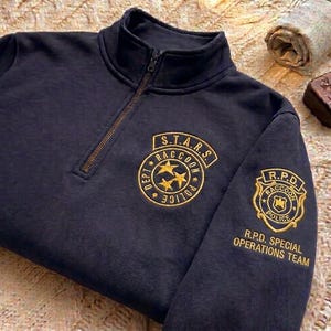 Może przedstawiać: Granatowa bluza z zamkiem błyskawicznym i złotymi haftowanymi naszywkami. Naszywki zawierają tekst "S.T.A.R.S. Raccoon Police Dept" i "R.P.D. Special Operations Team". Bluza ma wysoki kołnierz i brązowy zamek błyskawiczny.