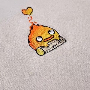Op de afbeelding: Geborduurde patch met een cartoon vuurduivel met een lachend gezicht, oranje vlammen en een geel hart. De patch is gestikt op een lichtbeige stof, wat de gedetailleerde borduurwerk laat zien.