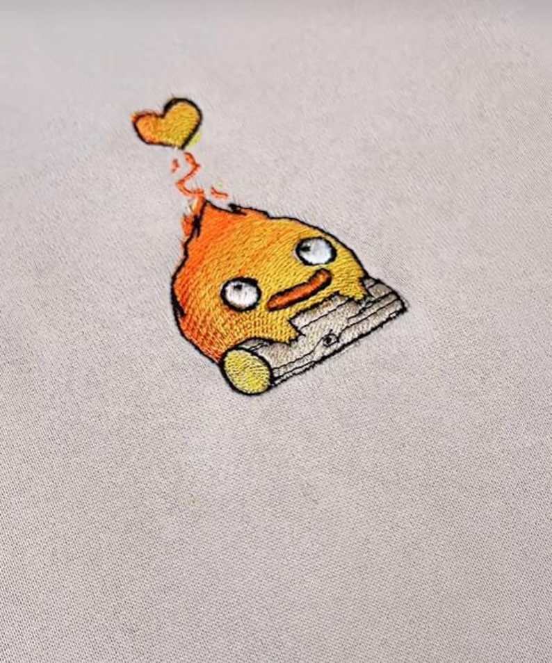 Op de afbeelding: Geborduurde patch met een cartoon vuurduivel met een lachend gezicht, oranje vlammen en een geel hart. De patch is gestikt op een lichtbeige stof, wat de gedetailleerde borduurwerk laat zien.