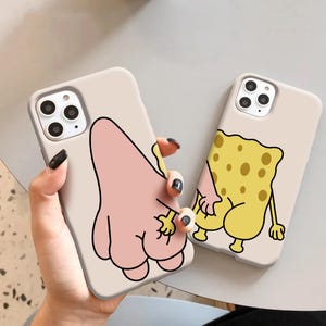 May include: Two beige phone cases with cartoon character designs. One case features a pink starfish character, and the other has a yellow sponge character. Both cases have black outlines and are held by a person.