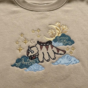May include: A beige crewneck sweatshirt featuring an embroidered design of a six-legged bison creature flying among clouds. The embroidery includes a yellow crescent moon, gold stars, and dark teal and light blue swirling cloud patterns.