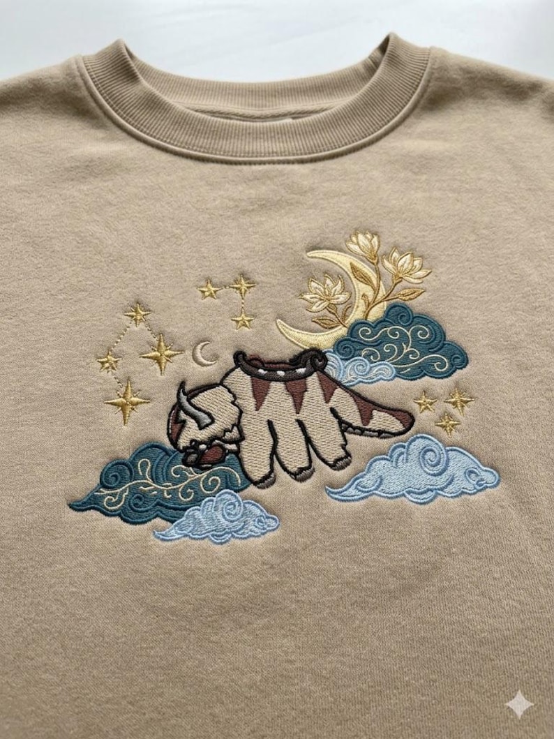 May include: A beige crewneck sweatshirt featuring an embroidered design of a six-legged bison creature flying among clouds. The embroidery includes a yellow crescent moon, gold stars, and dark teal and light blue swirling cloud patterns.