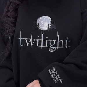 May include: Black sweatshirt with white embroidered design. Features the word "twilight" in a stylized font, a moon and trees graphic, and text on the sleeve that reads "And so the lion fell in love with the lamb."
