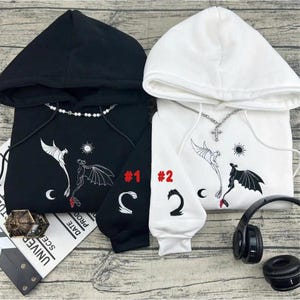 May include: Two hooded sweatshirts, one black and one white, with dragon and sun designs. The black sweatshirt has the number "1" and a crescent moon on the sleeve. The white sweatshirt has the number "2" and a crescent moon on the sleeve.