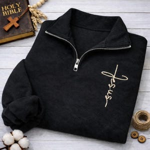 Könnte beinhalten: Schwarzes Sweatshirt mit silbernem Reißverschluss und dem Wort "Jesus" in goldener Schrift gestickt. Das Sweatshirt hat einen hohen Kragen und lange Ärmel. Das Bild enthält auch eine Bibel, ein Holzkreuz und andere religiöse Gegenstände.