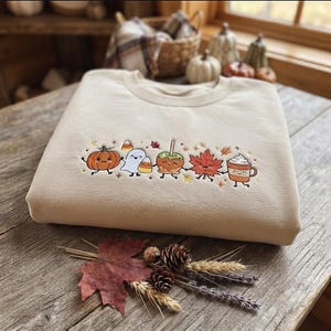 Könnte beinhalten: Ein beigefarbenes Sweatshirt mit Rundhalsausschnitt und einer zentralen horizontalen Stickerei von niedlichen, lächelnden Herbstmotiven, darunter ein Kürbis, ein Geist, Zuckermais, ein Liebesapfel, ein Ahornblatt und ein Becher Kakao.
