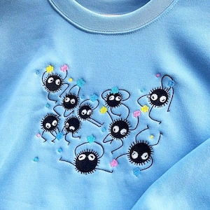 May include: Pale blue sweatshirt with an embroidered design of black, round, cartoon-like creatures with white eyes and small limbs. The design includes colourful star-shaped accents. The sweatshirt is hanging on a wooden hanger.