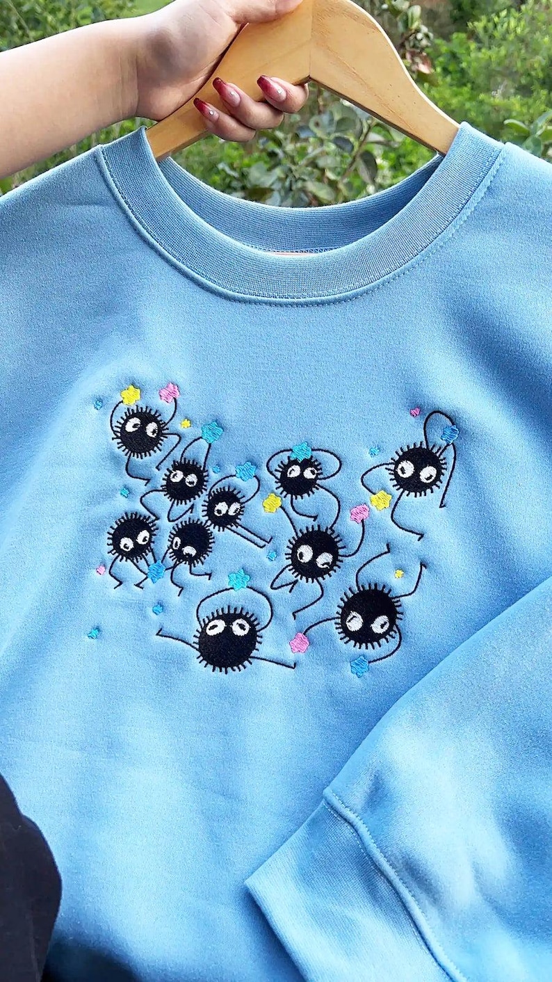 May include: Pale blue sweatshirt with an embroidered design of black, round, cartoon-like creatures with white eyes and small limbs. The design includes colourful star-shaped accents. The sweatshirt is hanging on a wooden hanger.