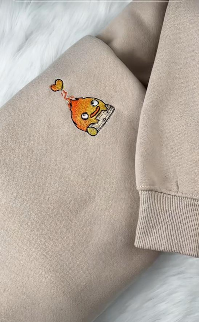 Op de afbeelding: Een beige sweatshirt met een geborduurd cartoonfiguur. Het figuur is oranje en geel met een lachend gezicht en een klein hartje erboven. De sweatshirt heeft een geribde manchet.