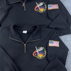 May include: Two black quarter-zip sweatshirts. Each features a "Project Hail Mary" patch with a rocket and sun design, and an American flag patch on the sleeve. The sweatshirts have gold zippers.