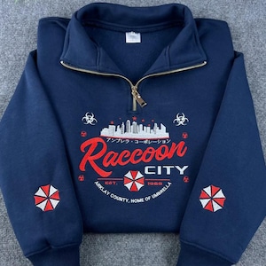 Può includere: Felpa blu navy con collo con zip. Presenta la scritta "Raccoon City" in rosso, una grafica del paesaggio urbano e il logo Umbrella Corporation. Include il testo "Arklay County, Home of Umbrella" e l'anno "1968".