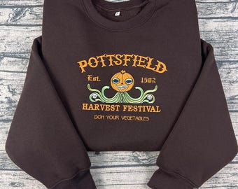 May include: A dark brown sweatshirt with the text "Pottsfield Harvest Festival" in orange and green. The design includes a smiling pumpkin character and the phrase "Don Your Vegetables."