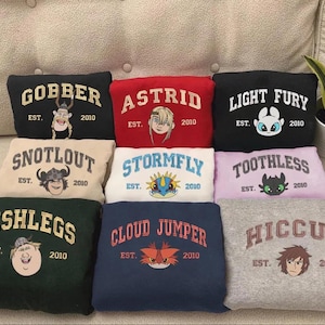 Op de afbeelding: Een verzameling opgevouwen sweatshirts in diverse kleuren, elk met een personagenaam en afbeelding uit de "How to Train Your Dragon" franchise. De namen zijn "Gobber", "Astrid", "Light Fury", "Snotlout", "Stormfly", "Toothless", "Fishlegs", "Cloud Jumper" en "Hiccup".