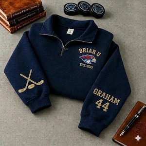 Peut inclure: Un sweat-shirt bleu marine à fermeture éclair quart, orné d'une broderie dorée indiquant Briar U Est. 2015 avec un logo d'oiseau. La manche droite présente deux crosses de hockey croisées et un palet, tandis que la gauche porte le nom Graham et 44.