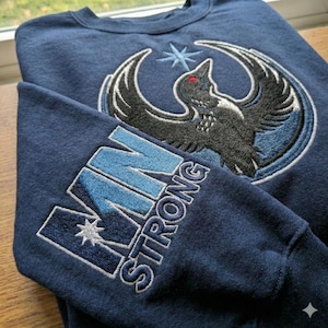 May include: Navy blue sweatshirt featuring an embroidered raven design and the text "VAN STRONG" on the sleeve. The raven is black and grey with a red eye, set against a blue circle with a star.