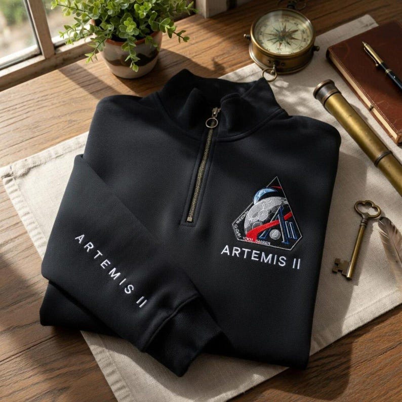May include: Black quarter-zip sweatshirt with the text "ARTEMIS II" on the sleeve and a triangular patch featuring a space-themed design. The zipper is gold-colored.