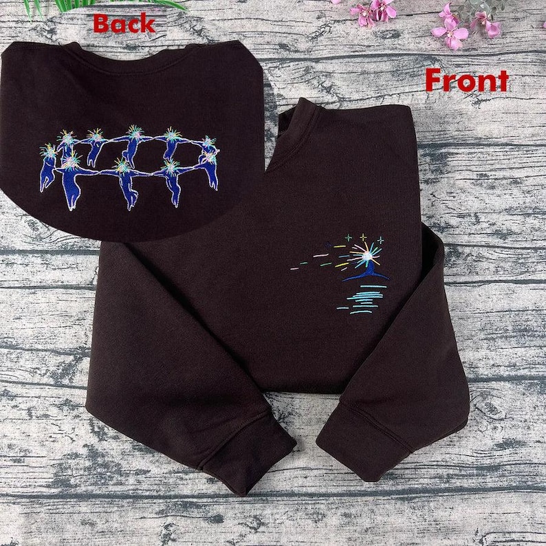 May include: A dark brown sweatshirt with embroidered designs. The front features a colourful sunburst design over a blue water line. The back has a circular design of blue figures holding hands. The sweatshirt has long sleeves.