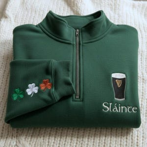 May include: A dark green fleece pullover with a quarter-zip closure. The left sleeve cuff features embroidered shamrocks in green, white, and orange. The right chest has an embroidered pint of stout and the word "Sl&aacute;inte" in white.