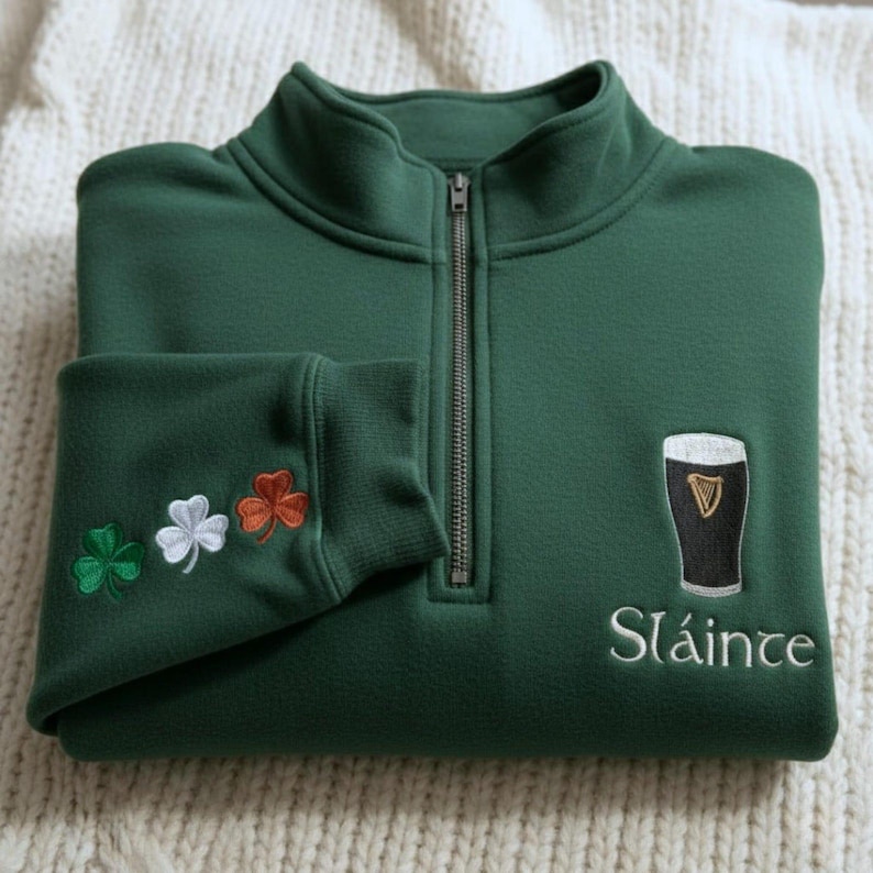 May include: A dark green fleece pullover with a quarter-zip closure. The left sleeve cuff features embroidered shamrocks in green, white, and orange. The right chest has an embroidered pint of stout and the word "Sl&aacute;inte" in white.