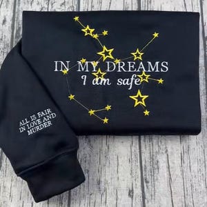 May include: Black sweatshirt with embroidered gold stars and the text "IN MY DREAMS I am safe." The sleeve features the text "ALL IS FAIR IN LOVE AND MURDER." The sweatshirt is folded on a wooden surface.