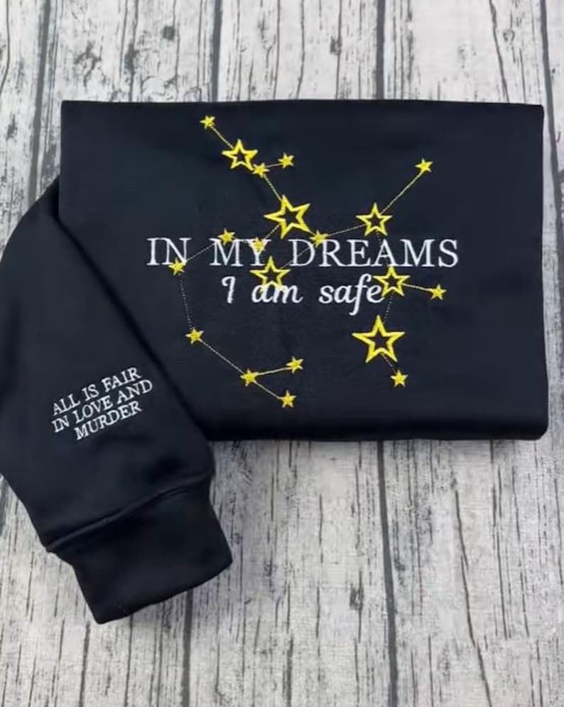 May include: Black sweatshirt with embroidered gold stars and the text "IN MY DREAMS I am safe." The sleeve features the text "ALL IS FAIR IN LOVE AND MURDER." The sweatshirt is folded on a wooden surface.