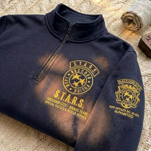 Może przedstawiać: Granatowa bluza z zamkiem błyskawicznym i złotymi haftowanymi naszywkami. Naszywki zawierają napisy "S.T.A.R.S." i "Raccoon City Police". Bluza ma postarzany wygląd.