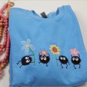 May include: A light blue sweatshirt featuring an embroidered design of four black, round characters holding colorful flowers. The flowers include a white, yellow, and pink design. The sweatshirt is made of a soft material.