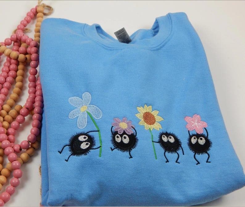 May include: A light blue sweatshirt featuring an embroidered design of four black, round characters holding colorful flowers. The flowers include a white, yellow, and pink design. The sweatshirt is made of a soft material.