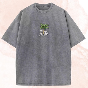 May include: A gray, short-sleeved t-shirt with a crew neck. The shirt features an embroidered design of a palm tree, two chairs, and a fruit. The shirt has a faded, vintage look.
