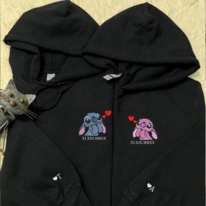 Puede incluir: Dos sudaderas con capucha negras con personajes de dibujos animados bordados, una azul y otra rosa, cada una con detalles de corazones rojos. Las sudaderas tienen fechas a juego en n&uacute;meros romanos y una letra y un coraz&oacute;n en las mangas.