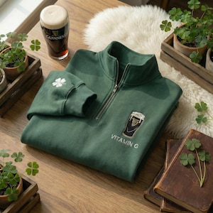 Pode incluir: Camisola verde-escura com fecho de correr de um quarto de gola, com um desenho de copo de Guinness e o texto "Vitamin G". Um trevo branco bordado na manga. Um copo de Guinness e plantas de trevo tamb&eacute;m s&atilde;o vis&iacute;veis.