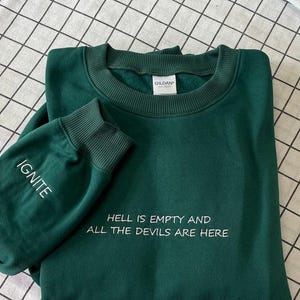 May include: A dark green sweatshirt with the text "HELL IS EMPTY AND ALL THE DEVILS ARE HERE" in white. The left sleeve has the word "IGNITE" in white. The sweatshirt is folded on a checkered surface.