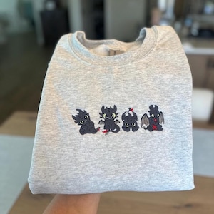 May include: A light gray sweatshirt featuring an embroidered design of four cartoon dragons in black, green, and red. The dragons are arranged in a horizontal row across the chest of the sweatshirt. The sweatshirt is folded.