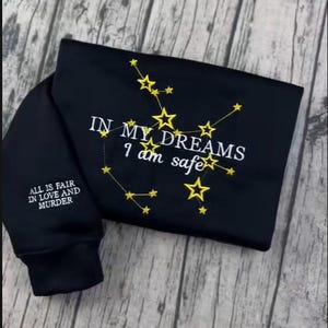 May include: Black sweatshirt with embroidered gold stars forming constellations. The text "IN MY DREAMS I am safe" is embroidered on the front. The sleeve reads "ALL IS FAIR IN LOVE AND MURDER".