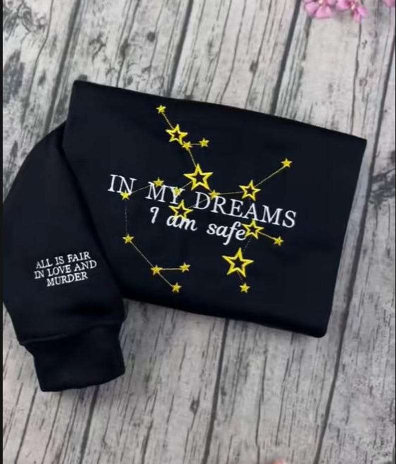 May include: Black sweatshirt with embroidered gold stars forming constellations. The text "IN MY DREAMS I am safe" is embroidered on the front. The sleeve reads "ALL IS FAIR IN LOVE AND MURDER".