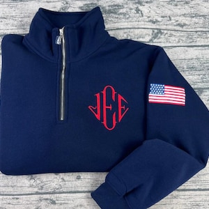 May include: Navy blue pullover sweatshirt with a quarter-zip closure and a mock neck. Features a red monogram on the chest and an American flag patch on the sleeve. The sweatshirt is made of a soft, comfortable material.