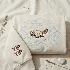 May include: A cream-colored sweatshirt featuring an embroidered brown and white flying bison surrounded by light blue stitched clouds. The sleeve displays the brown embroidered text YIP YIP. The design is centered on the front of the garment.