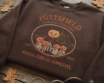 May include: A dark brown crewneck sweatshirt featuring an embroidered design of five pumpkin-headed figures. The text reads POTTSFIELD HARVEST FESTIVAL EST. 1502 and YOU'LL JOIN US SOMEDAY. The graphic is enclosed in a rope-style oval border.